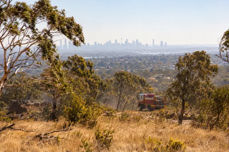 Scena editoriale contestuale di un paesaggio arido nella regione di Sydney durante il caldo anomalo, con un mezzo antincendio in prevenzione